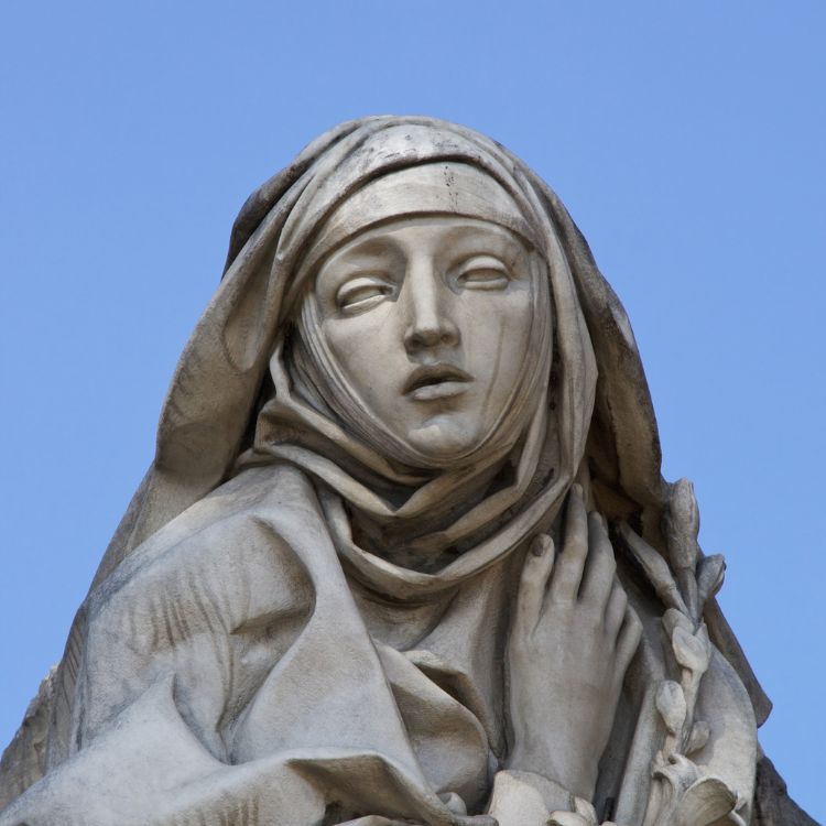 Estátua de Santa Catarina de Siena feita em mármore claro, apresentando expressão serena e olhar voltado para cima. Santa utiliza um véu e segura as vestes com uma das mãos. Fundo azul claro representa o céu limpo.