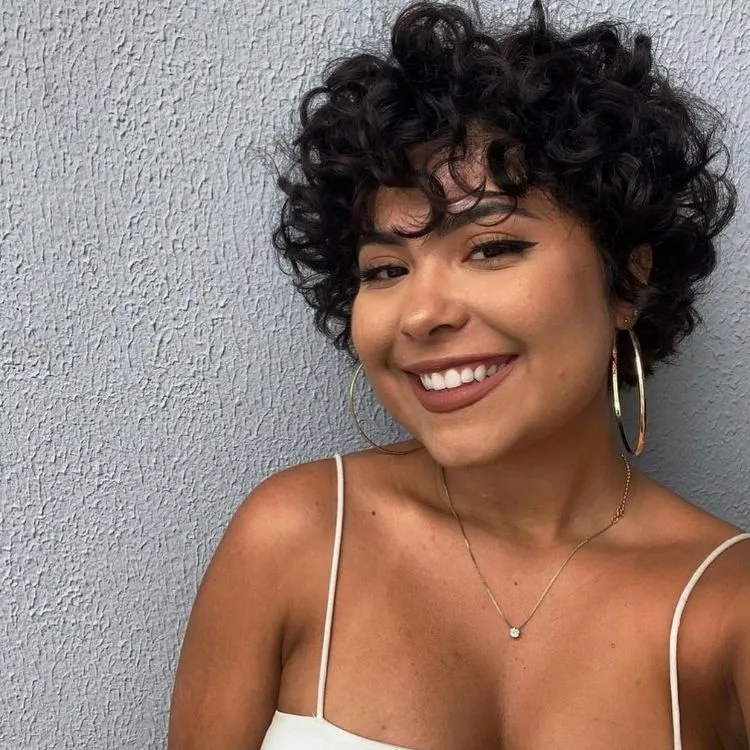Uma mulher de pele morena com um corte de cabelo bem curto e cacheado, ela usa uma blusa de alças brancas