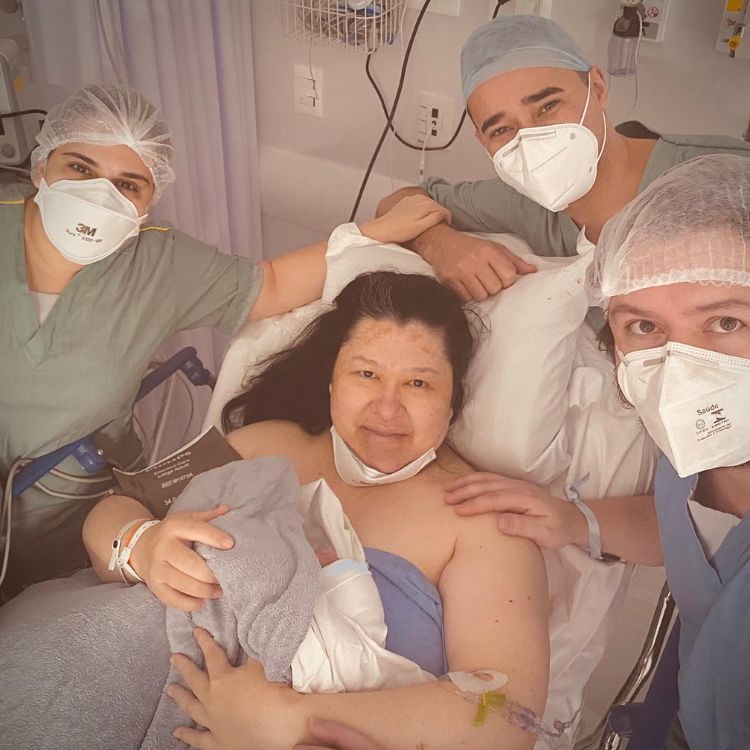 Paloma Sato na maca com bebê no colo, Atila e médicos de roupa cirúrgica, touca e máscara