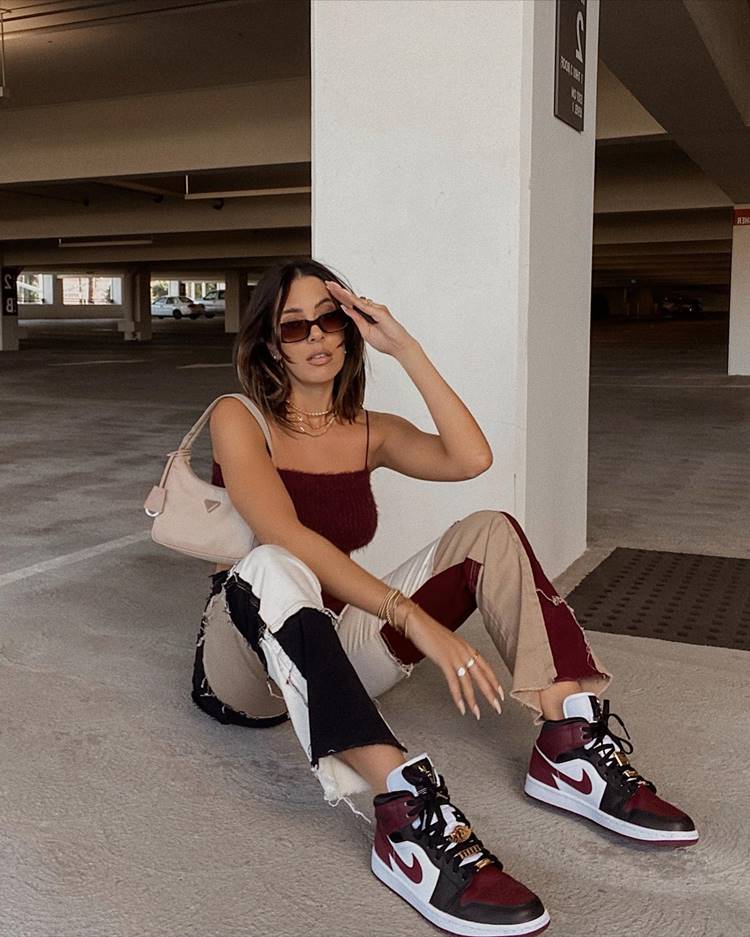 Garota sentada no chão usando regata de pelinho, calça patchwork nas cores bege, vinho e branco, tênis da Nike e bolsa, em estacionamento de prédio.
