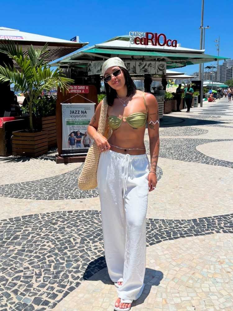 Mulher de pele clara em orla da praia usando biquini, calça branca, lenço verde, bolsa de palha e bracelete