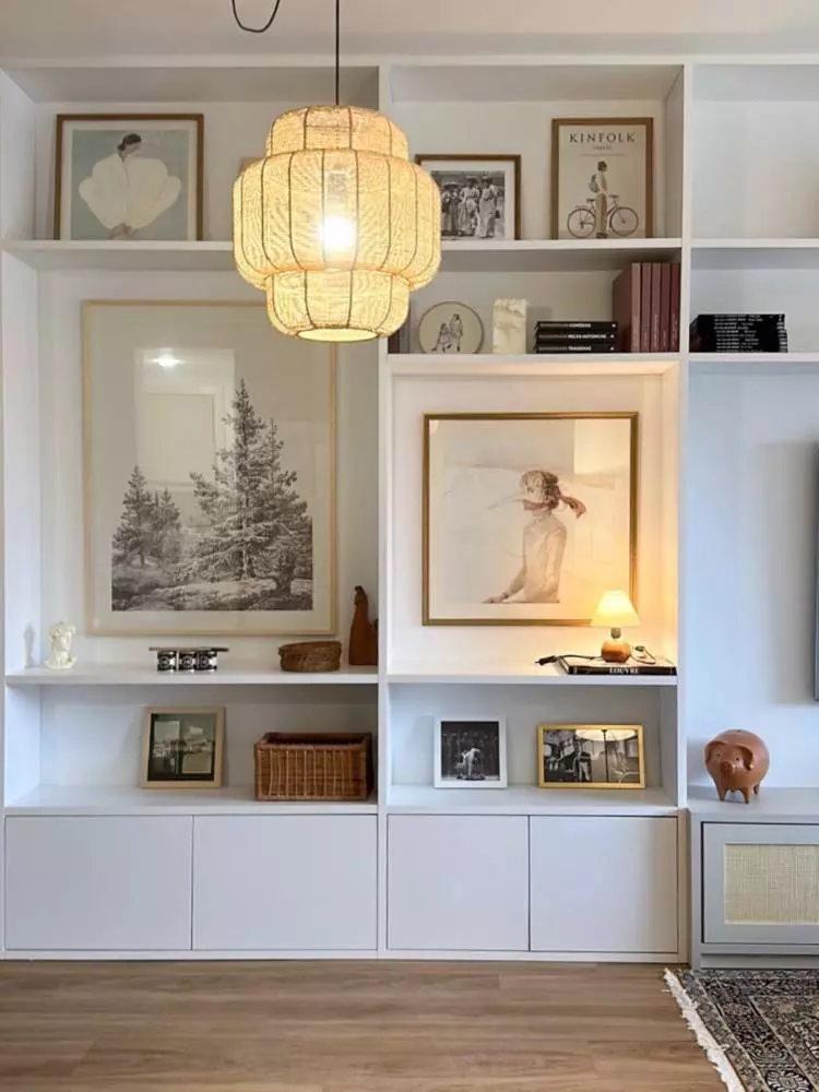 Estante de madeira com nichos abertos em cima e fechados em baixo, em branco. Nela, há uma luminária acesa, diversos quadros de f=diferentes tamanhos, livros, porta-retratos e outros itens decorativos. 