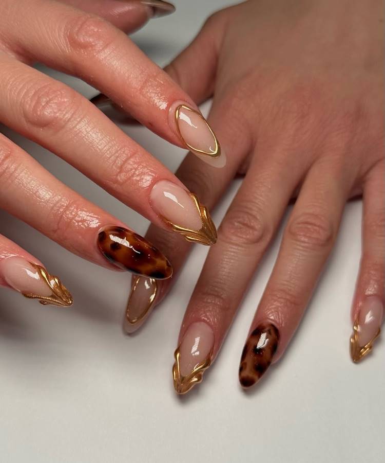 A imagem mostra mãos com unhas em formato alongado e acabamento sofisticado. Algumas têm design tartaruga em tons marrons, enquanto outras exibem esmalte nude com bordas douradas metálicas. O fundo claro destaca os detalhes elegantes e modernos da nail art.