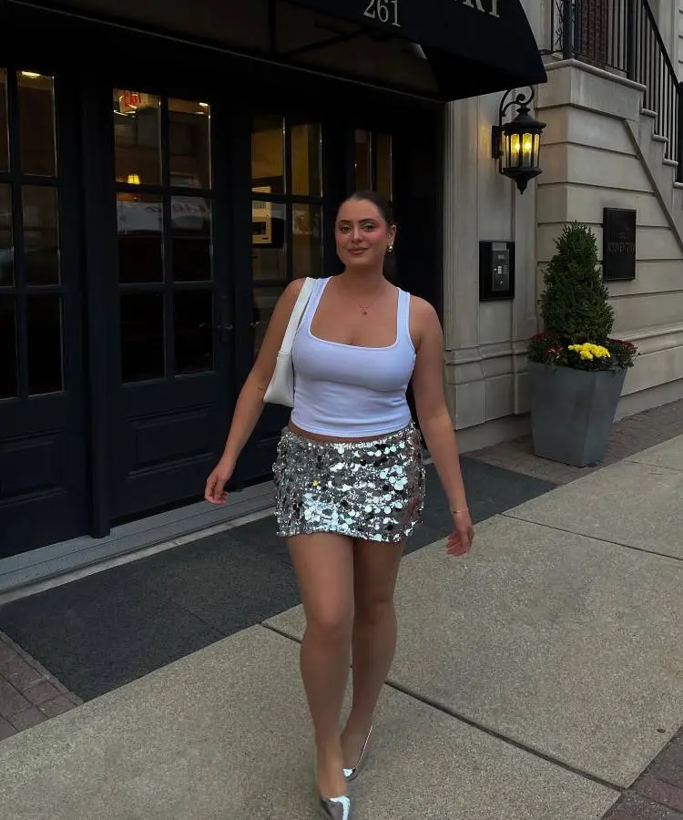 A mulher caminha confiante em uma calçada elegante, vestindo uma blusa branca básica e uma saia curta prateada com paetês brilhantes. Completa o look com sapatos metálicos, bolsa branca e brincos delicados, destacando um estilo sofisticado e moderno.
