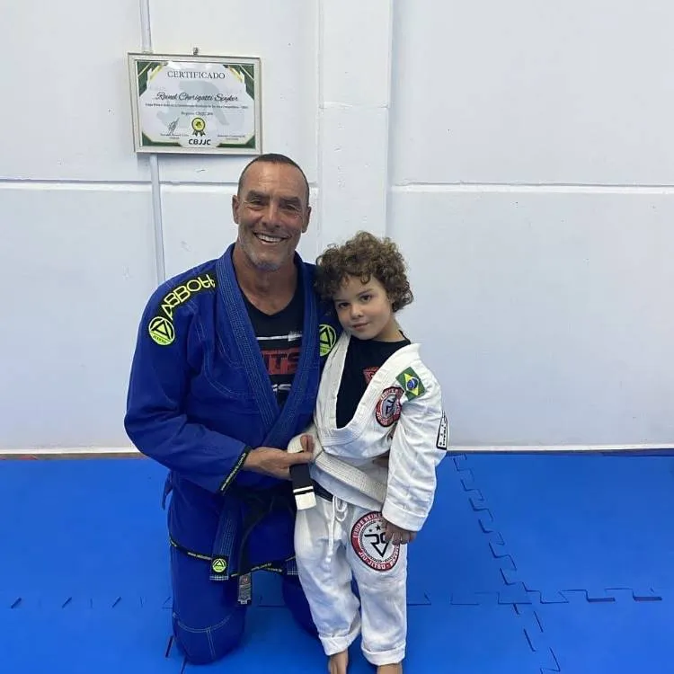 Um homem sorridente de meia-idade, vestindo um kimono de jiu-jítsu azul, posa ajoelhado ao lado de uma criança com cabelos cacheados. A criança, vestindo um kimono branco com a bandeira do Brasil, sorri levemente. Ambos estão em um tatame azul com uma parede branca ao fundo.

