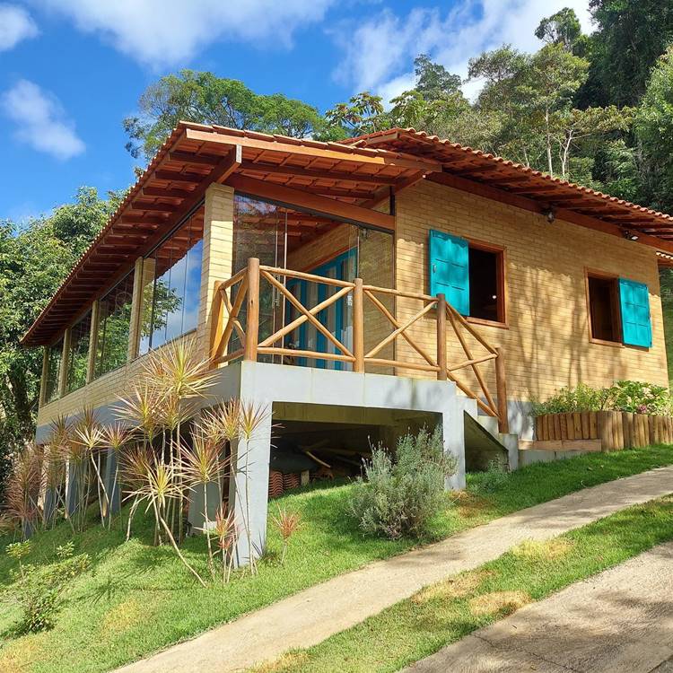 Imagem de fachada de casa simples em terreno inclinado. A casa possui acabamento em tijolinhos, janelas e portas azuis. Uma escada leva para a entrada principal. Corrimão feito em madeira