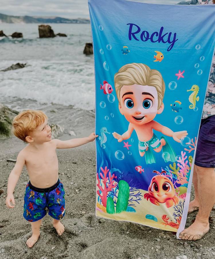 Um menino pequeno com shorts de banho coloridos, sorri ao lado de uma toalha de praia azul e personalizada com o nome "Rocky". A toalha mostra um personagem de desenho animado no fundo do mar.

