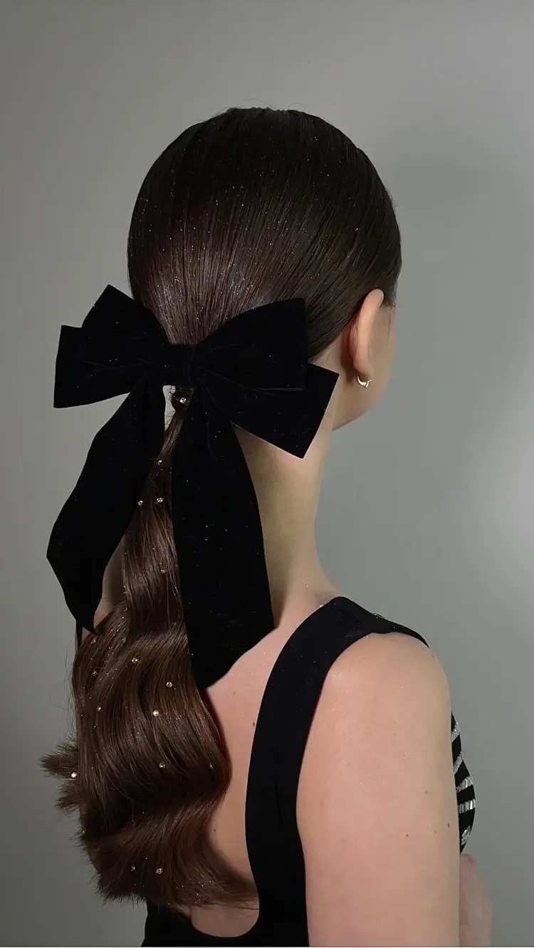Mulher de pele clara de costas usando vestido preto e cabelo castanho em rabo baixo com glitter, strass e laço preto