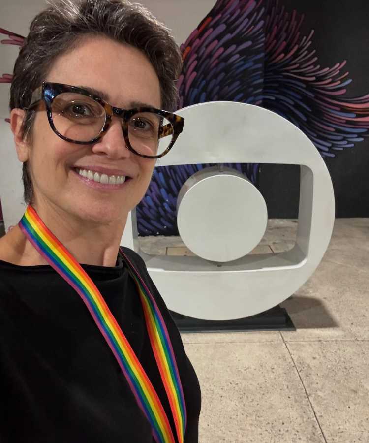 Sandra Annenberg sorridente com óculos de armação tartaruga e cabelo curto posa em frente ao logotipo da Globo. Usando blusa preta e faixa arco-íris, ela demonstra confiança e é frequentemente citada entre os famosos que não envelhecem.