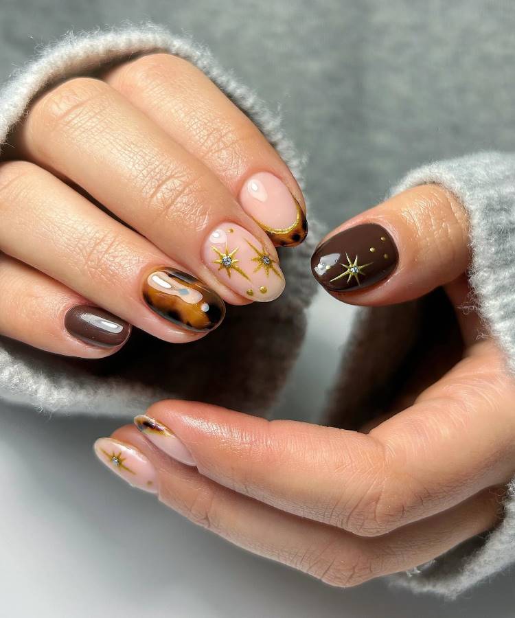 Unhas de gel decoradas com esmaltes em tons marrons e nude, detalhes marmorizados, estrelas douradas e acabamento brilhante. O design moderno e elegante combina texturas e cores sofisticadas, criando um estilo único e envolvente para destacar as mãos.