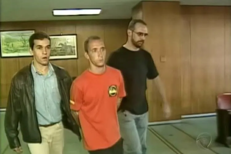 Cristian Cravinhos usando uma blusa vermelha sendo conduzido pela delegacia por dois homens, um de blusa preta e outro de blusa azul.