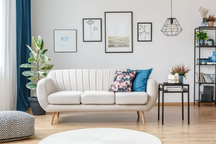 Sala com sofá off white arredondado, almofada floral, quadros fofos, tapete branco redondo e planta