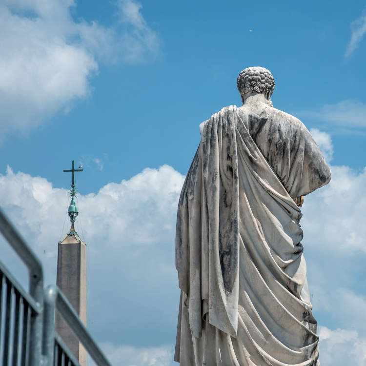 Foto de estátua de São Pedro de costas, ao lado de igreja e diante de um céu azul
