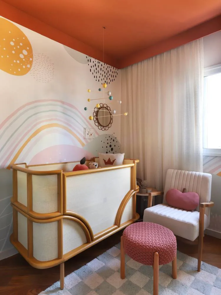 Esta é uma imagem de um quarto de bebê vibrante com um teto colorido laranja. O berço de madeira clara com tela de vime se destaca contra a parede com um mural de arco-íris e bolinhas. Um pufe rosa adiciona um toque de cor, completando o ambiente lúdico.

