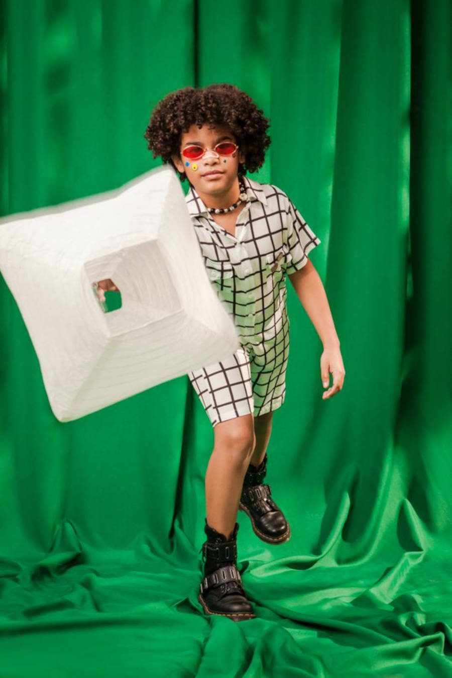 Foto de criança utilizando um conjunto xadrez e arremessando um balão quadrado para a câmera. O fundo da imagem é verde.