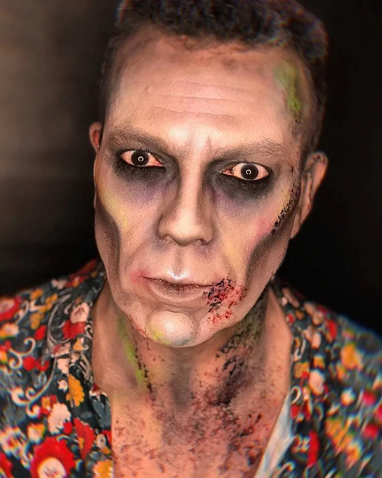 Homem de pele clara com make de zumbi, com manchas pretas, manchas verdes e sangue falso 