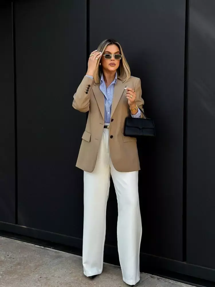 Mulher loira de pele clara usando blazer bege, camisa listrada azul e branca e calça social branca 