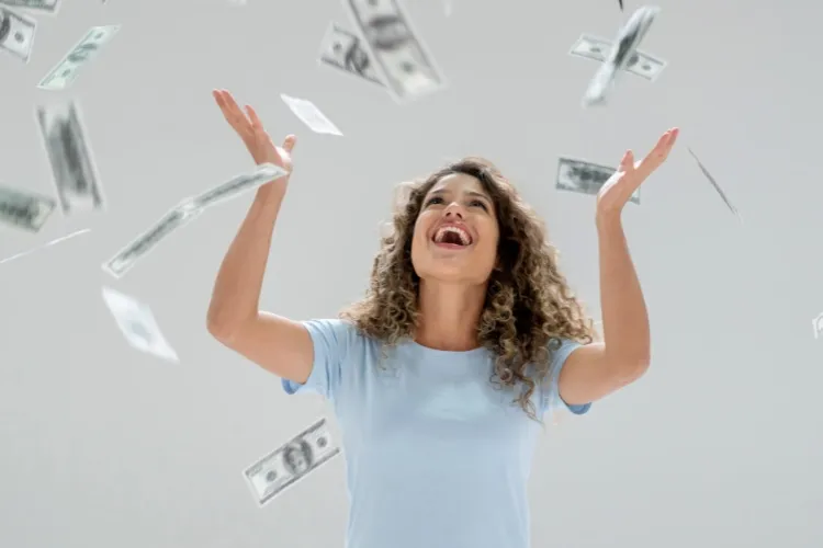 Mulher de pele morena usando blusa azul clara e olhando feliz para cima, enquanto cédulas de dinheiro caem