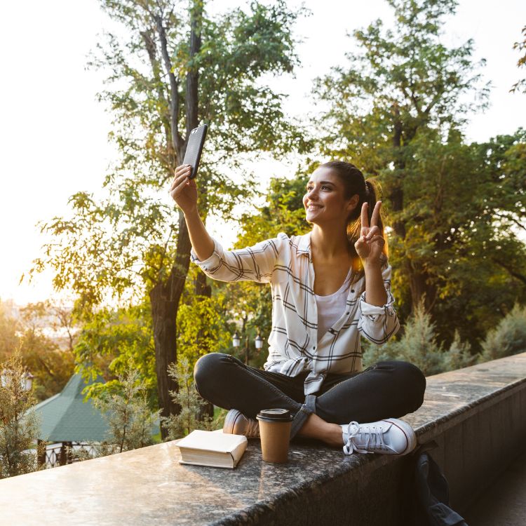 Jovem influencer sentada ao ar livre, em um ambiente arborizado, tirando uma selfie enquanto faz o gesto de paz. Ao lado, um livro e café. Representa o que fazem os influencers: criar conteúdo autêntico e engajar seguidores.