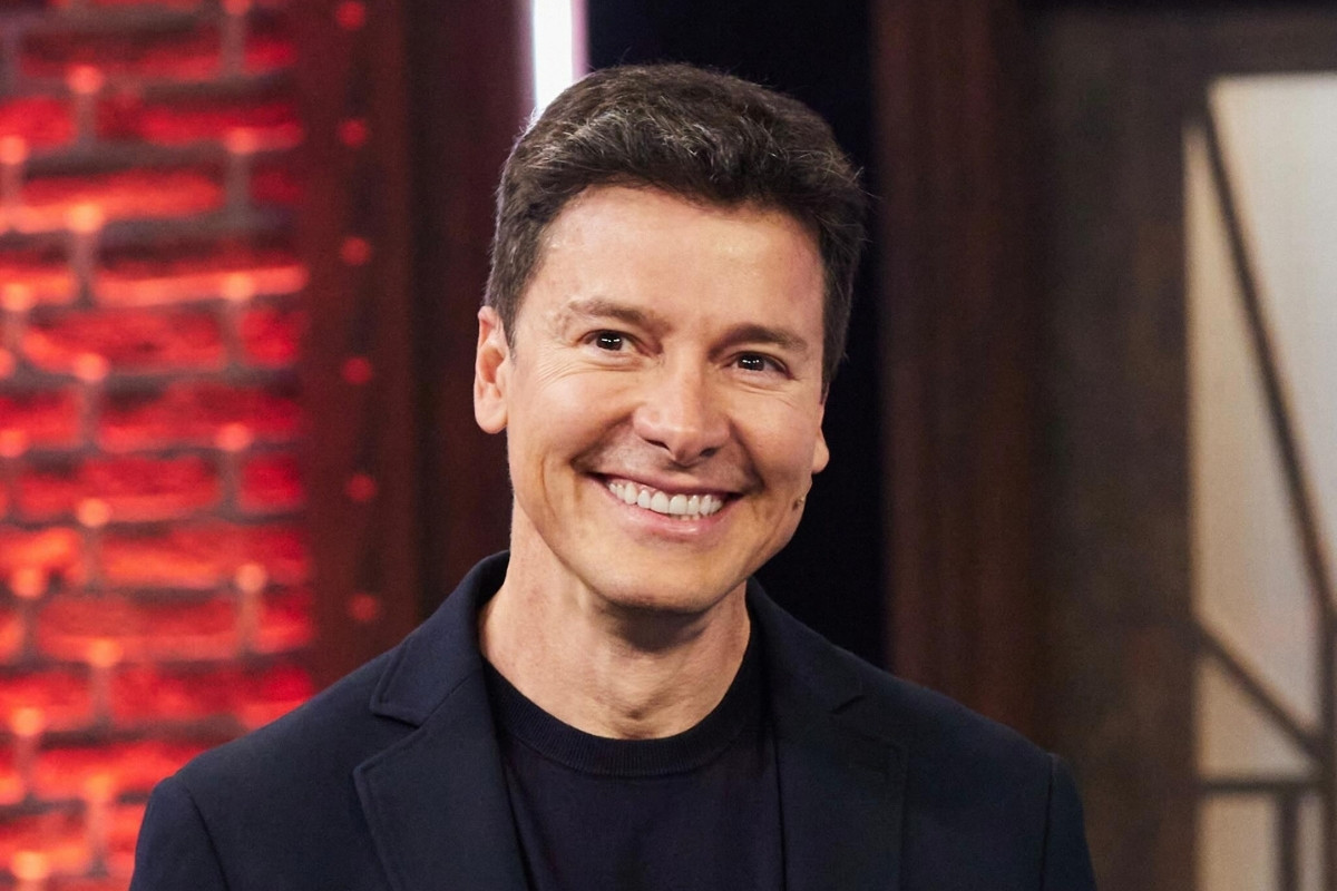Close-up de Rodrigo Faro, apresentador e ator brasileiro, sorrindo para a câmera em frente a um fundo de tijolos e luzes vermelhas. Ele veste um blazer azul marinho sobre uma camiseta preta, com cabelo castanho escuro e penteado.