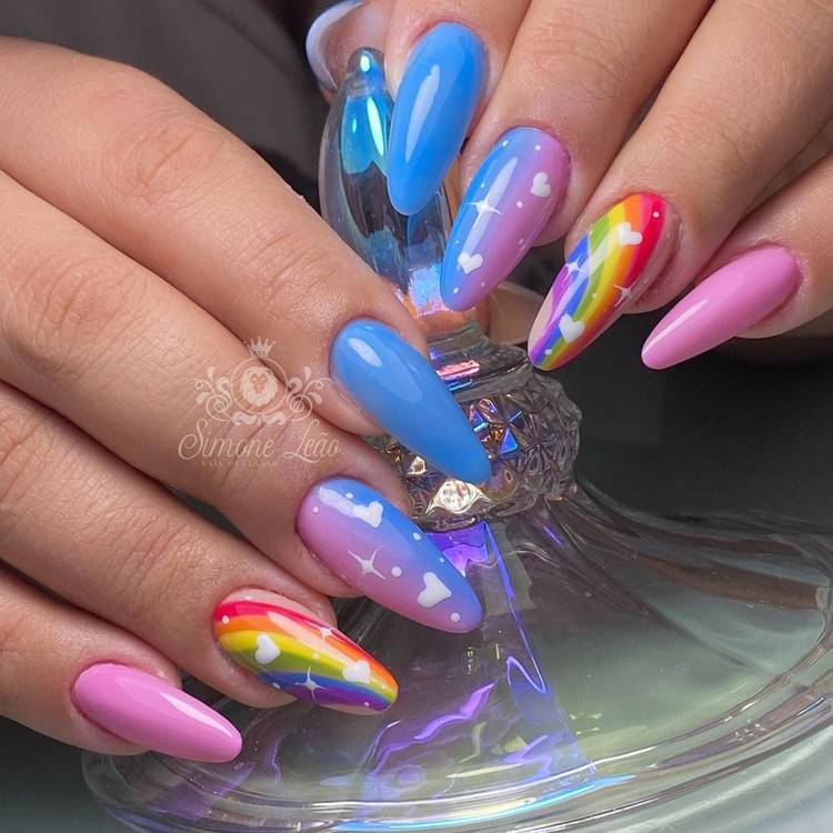 Mão com unhas decoradas coloridas: Polegar: não aparece na foto Indicador: azul Médio: rosa e azul mesclado com corações branco Anelar: arco-íris com corações brancos Mínimo rosa