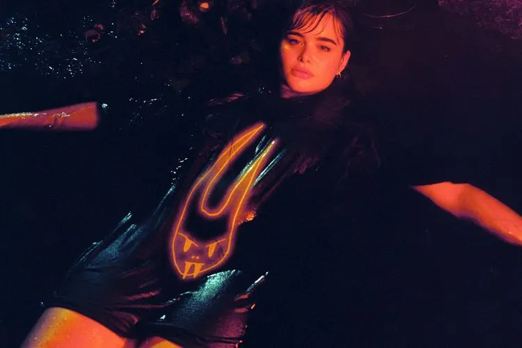 A atriz Barbie Ferreira posa deitada com uma camiseta preta com uma estampa de coelho, a blusa e o cabelo da atriz tem aspecto de molhado 