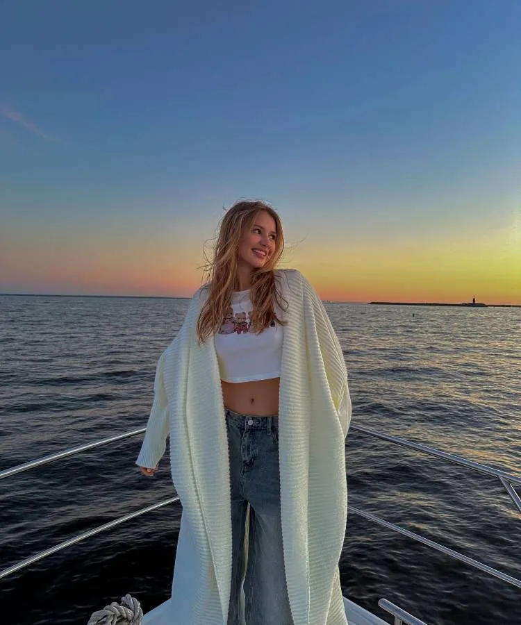 Uma mulher loira, vestindo um top branco e um casaco longo de tricô, está em um barco ao pôr do sol. O céu está colorido em tons de laranja e azul, e o mar calmo reflete a luz suave do crepúsculo.


