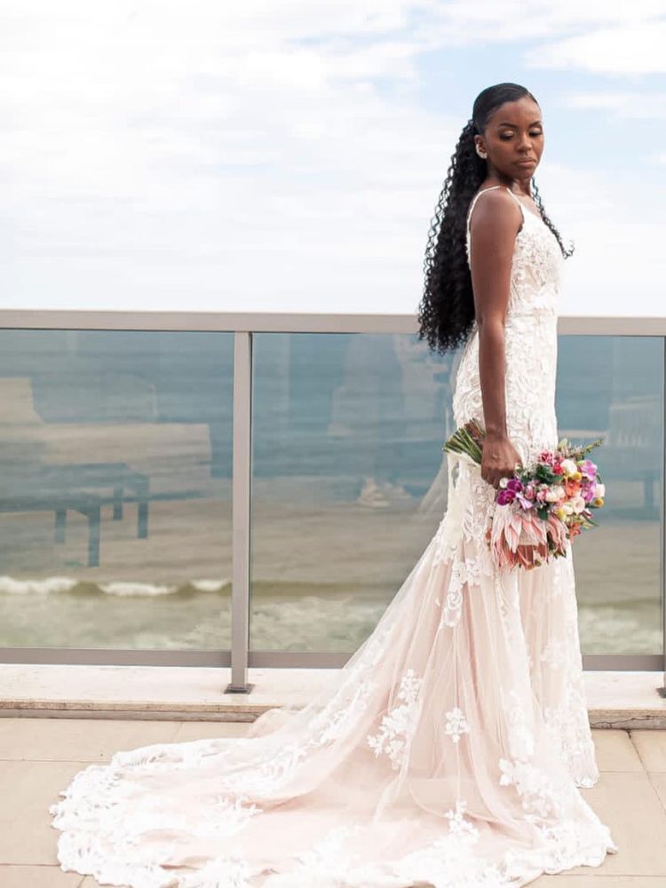 Noiva de pele negra usando vestido de alça, cabelo semi preso e segurandp buquê