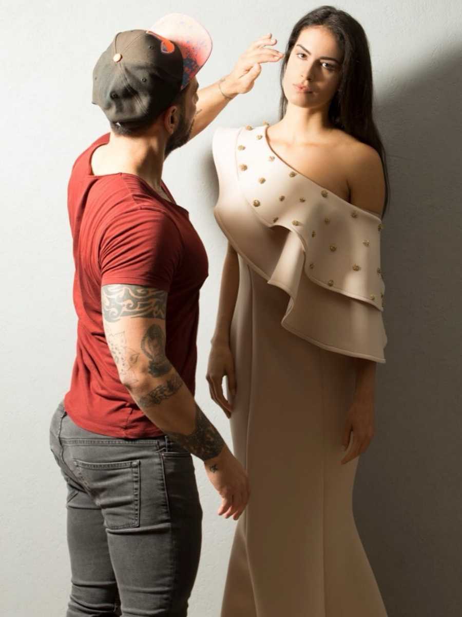 O designer vestindo modelo com vestido da sua marca AVVAA. Ele está tocando o cabelo dela enquanto ela está olhando fixamente para a câmera. Os dois estão em pé.