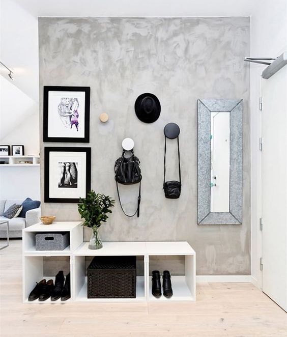 Hall de entrada com parede de cimento queimado, quadros, espelho, gancho para chapéu e bolsas e móvel branco modular com sapatos, caixas e vaso com planta