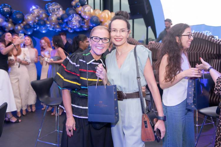 Duas mulheres posam sorridentes em frente a decoração com balões dourados e azuis em evento. Elas seguram sacola de presente. Ao fundo, várias pessoas conversam animadamente. Ambiente iluminado e festivo, com destaque para cores vivas e atmosfera elegante.