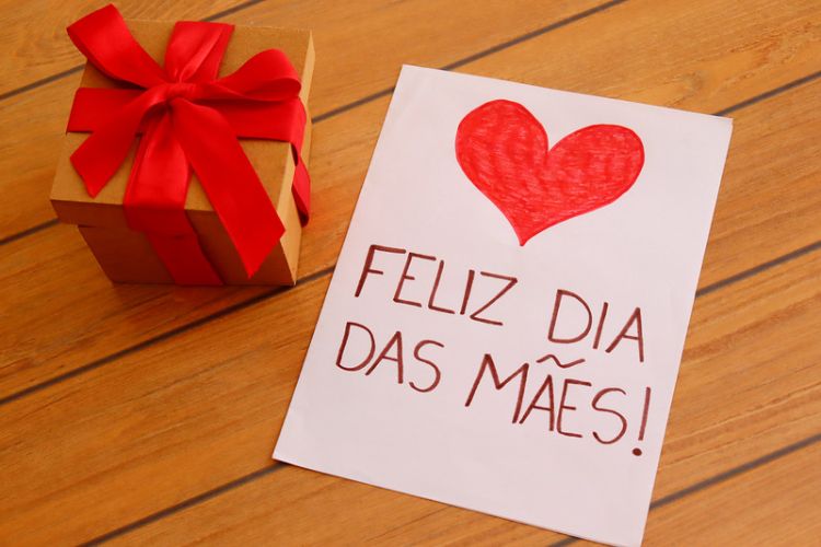 Mesa de madeira com presente e cartão escrito "feliz Dia das Mães" com coração