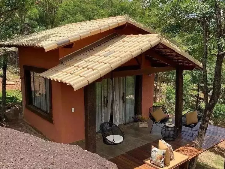 Fachada de casa pequena em área com árvores. Deck possui cadeiras em ferro e balanço