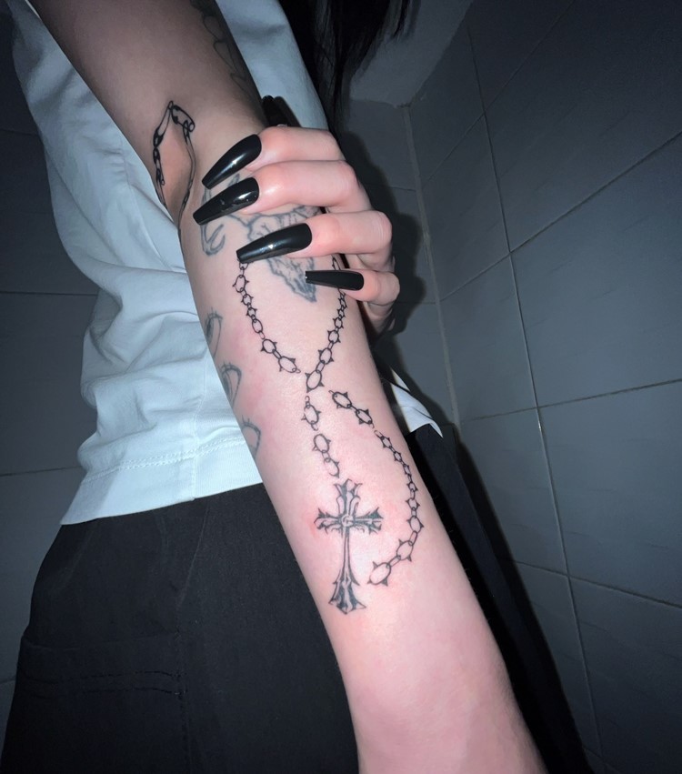 A foto mostra um braço com uma tatuagem de terço envolvendo o antebraço, destacando uma cruz detalhada na parte inferior. A pessoa veste uma camiseta branca e segura o braço, exibindo unhas longas e escuras. O fundo é composto por azulejos claros, criando um contraste com a pele e a tatuagem, ressaltando a arte religiosa.


