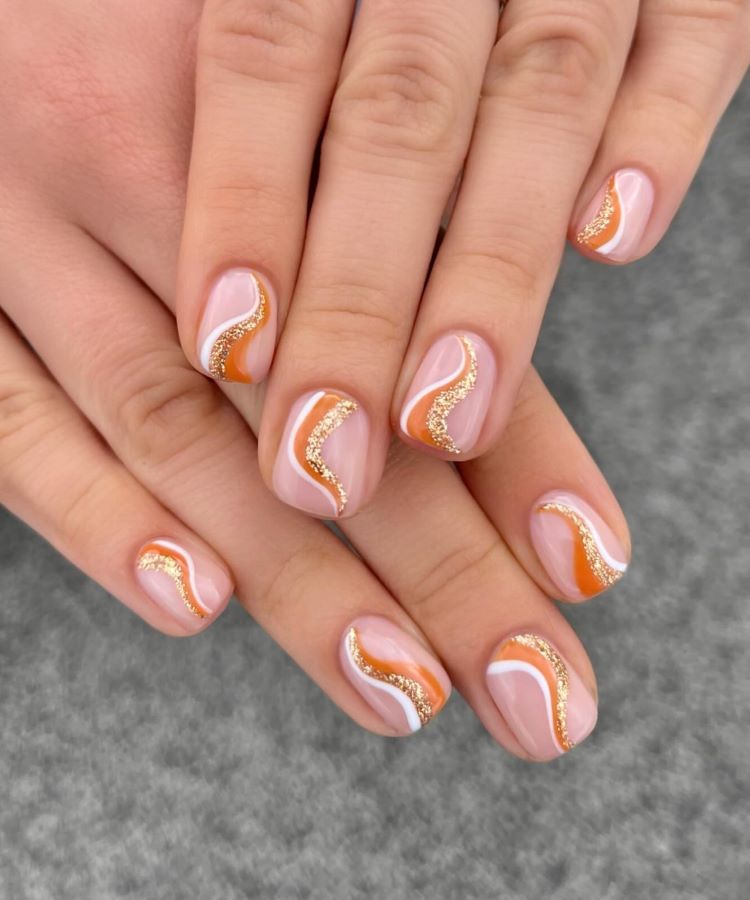 Mãos exibindo estilos de unhas curtas, decoradas com esmalte nude como base, detalhes ondulados em branco, laranja e glitter dourado. O design moderno combina elegância e criatividade. Fundo cinza texturizado realça os contrastes suaves e o acabamento impecável das unhas.