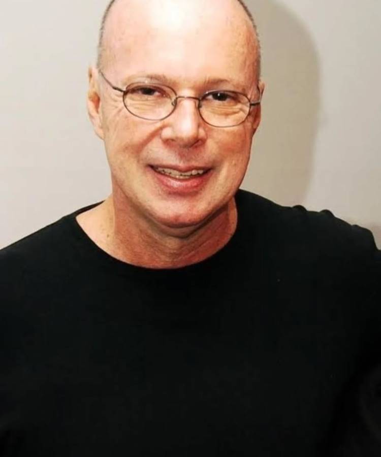 Close-up de um homem branco de meia-idade, careca, sorrindo gentilmente para a câmera. Ele usa óculos de armação fina de metal e uma camiseta preta básica. O fundo é liso e claro.