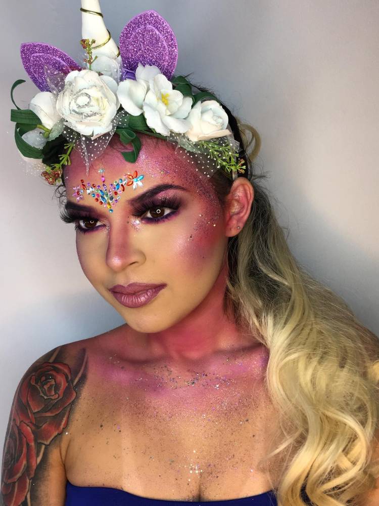 Mulher de pele clara e cabelo loiro usando tiara de unicórnio, muito brilho roxo no rosto e no colo, maquiagem roxa com cílios postiços e coração de cartela de adesivos. 