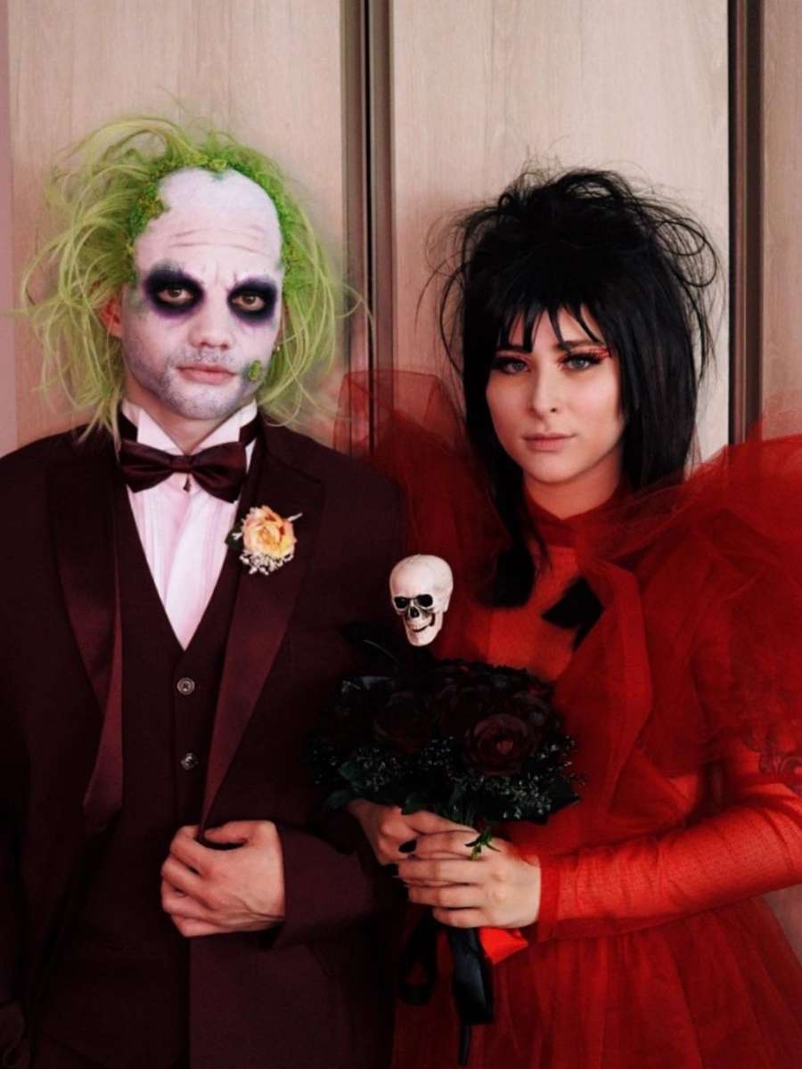 Imagem em fundo de guarda-roupa com Talita Kemy e Gustavo na frente. Eles vestem fantasia de Lydia Deetz e Beetlejuice, personagens do filme "Os Fantasmas se divertem" (1988). Gustavo está usando terno de cor vinho, com o rosto pintado de branco e peruca verde. Talita veste vestido vermelho, peruca preta bagunçada e segura buquê de flores preto com caveira.