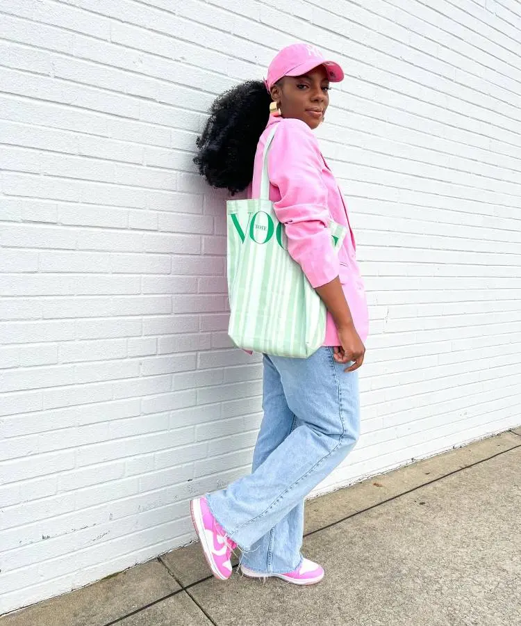 Uma mulher sorridente com um rabo de cavalo alto e um boné rosa claro, posa de lado vestindo um blazer rosa vibrante, jeans de lavagem clara e tênis rosa e branco. Ela carrega uma bolsa tote listrada verde e branca com a palavra "VOGUE".