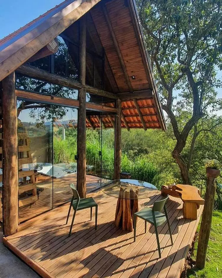 Imagem de fachada de chalé em área rural. Chalé de vidro e madeira, com deck, banco de madeira esculpido, mesa de vidro e cadeiras verdes