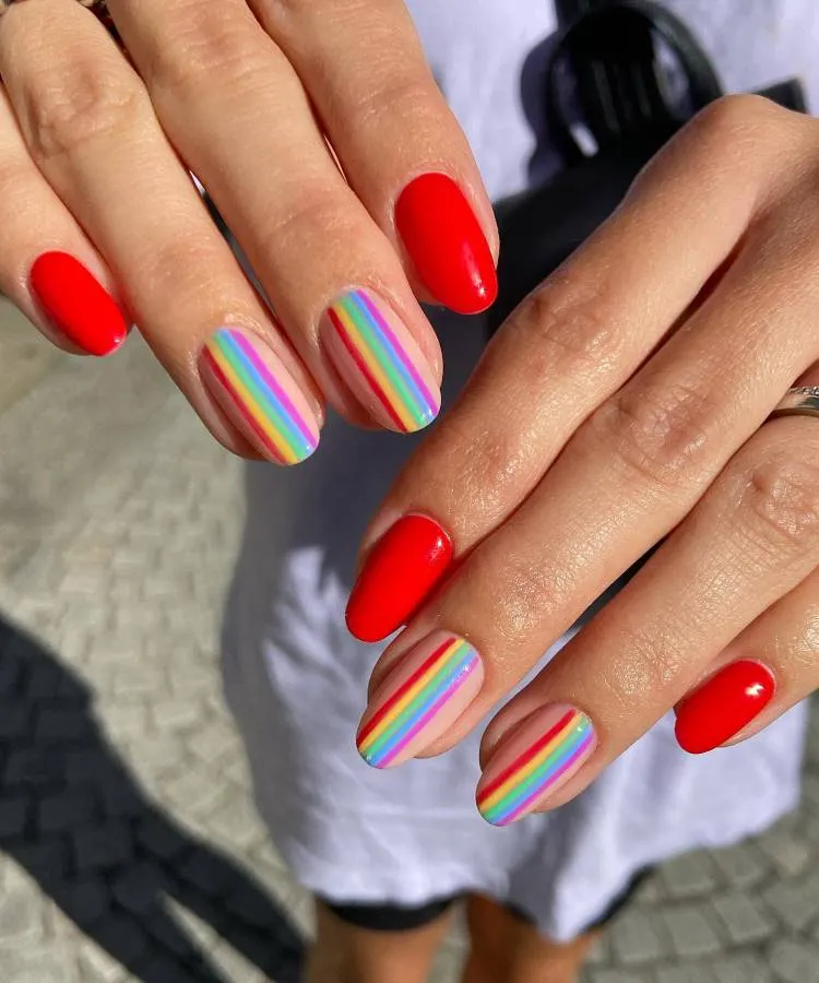 A imagem mostra um close-up da mão de uma pessoa exibindo suas unhas pintadas com esmalte. Há 8 unhas no total, cada uma com um design diferente. O dedo indicador e o dedo mindinho estão pintados com uma cor sólida vermelha. Os dedos médio e anelar têm um padrão listrado com várias cores, incluindo rosa, amarelo, verde, azul e possivelmente outras, criando um efeito de arco-íris. As unhas estão bem cuidadas, com formato oval e o esmalte tem um acabamento brilhante. 