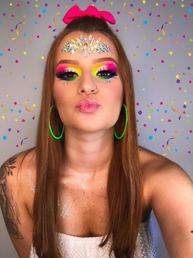 Mulher de pele clara e cabelo ruivo usando maquiagem colorida, strass na testa e brincos de argola verdes. 