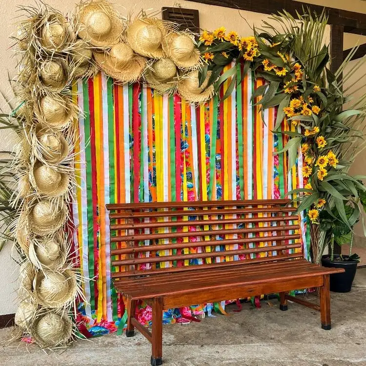 Um cenário de festa Junina com uma parede decorada com chapéus de palha, girassóis e fitas coloridas verticais. Um banco de madeira está posicionado à frente, perfeito para fotos.