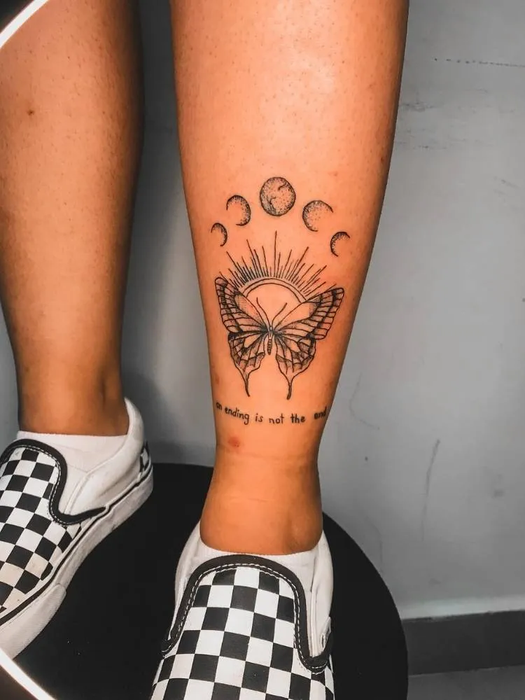 Tatuagem de Borboleta cheia de detalhes, sombreamento, traços finos e mais grossos. Ela acompanha  a frase "An ending is not the end" e as fases da lua acima. Foi feita na canela de uma mulher de pele clara que está usando um tênis quadriculado de branco e preto. 