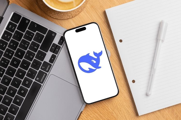 A imagem mostra um smartphone sobre um laptop, exibindo o logotipo azul de uma baleia estilizada em tela branca. Ao lado, há um caderno com folhas pautadas e uma caneta, além de uma xícara de café.