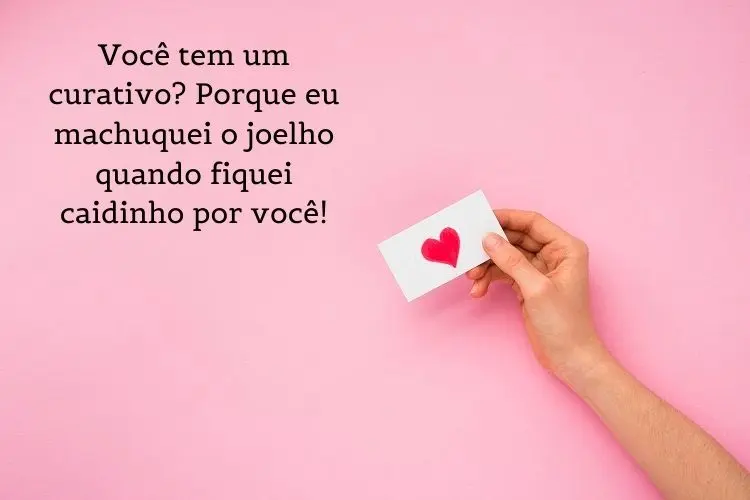 Cartão virtual de fundo rosa com pessoa de pele clara segurando papel com coração vermelho e frase 'Você tem um curativo? Porque eu machuquei o joelho quando fiquei caidinho por você!"