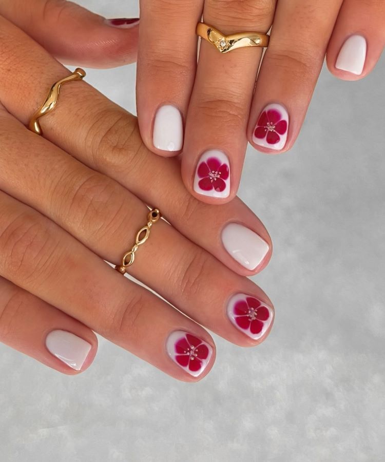 Mãos com estilos de unhas curtas, pintadas em branco clássico, destacando-se duas unhas decoradas com flores vermelhas vibrantes e miolo com brilho. Dedos adornados com anéis dourados delicados. Fundo cinza claro desfocado, destacando o visual minimalista e sofisticado.
