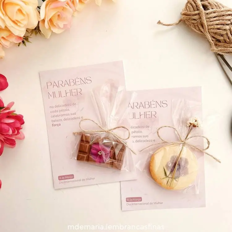 Imagem de lembrancinhas para o Dia Internacional da Mulher: cartão com a mensagem "Parabéns Mulher", acompanhado de chocolate decorado e biscoito personalizado, embalados com plástico transparente e laços de sisal. Fundo com flores artificiais e barbante decorativo.