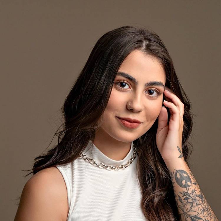 A imagem mostra uma mulher jovem com cabelo castanho comprido, sorrindo levemente. Ela veste uma blusa sem mangas branca e um colar prateado. Há uma tatuagem floral no braço esquerdo, e ela toca o rosto com a mão direita. Ela é uma das meninas da lista de blogueiros CLTs.

