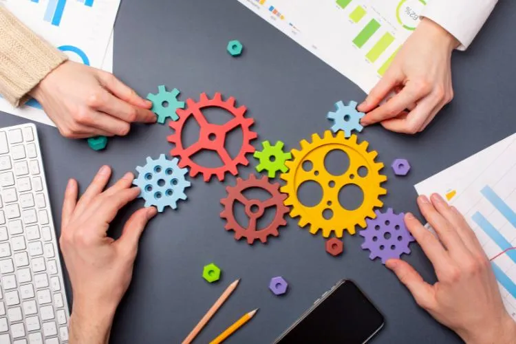 Quatro mãos ajustam engrenagens coloridas sobre uma mesa com papéis e gráficos, simbolizando trabalho em equipe e inovação. A imagem destaca colaboração estratégica para resolver problemas e "transforme crise em oportunidade" ao encontrar soluções criativas e eficientes.
